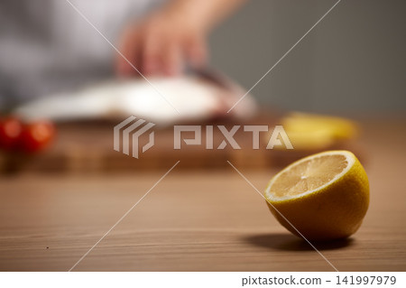 Lemon half in focus on a kitchen table, person preparing fresh fish and vegetables in blurred background, cooking seafood Lemon half in focus on a kitchen table, person preparing fresh fish and vegetables in blurred background, cooking seafood 141997979