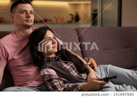 Young couple enjoying leisure time on a comfortable couch, watching tv with a remote control. Domestic life at home Young couple enjoying leisure time on a comfortable couch, watching tv with a remote control. Domestic life at home 141997980