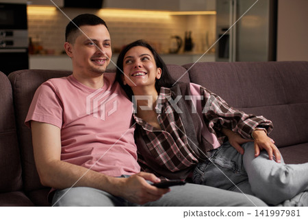 Happy couple enjoying a cozy evening controlling the tv with a remote, sharing leisure time on the sofa Happy couple enjoying a cozy evening controlling the tv with a remote, sharing leisure time on the sofa 141997981