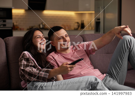 Happy couple relaxing on a sofa, watching tv and laughing together. woman holding remote control, pointing at screen Happy couple relaxing on a sofa, watching tv and laughing together. woman holding remote control, pointing at screen 141997983