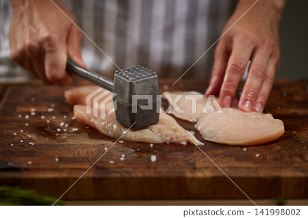 Chef's hands pounding and tenderizing raw chicken breast fillets with a meat mallet on a wooden cutting board in a kitchen Chef's hands pounding and tenderizing raw chicken breast fillets with a meat mallet on a wooden cutting board in a kitchen 141998002