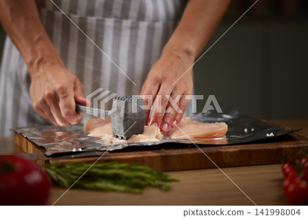 Cook tenderizing raw chicken meat with a metal mallet on a wooden board, preparing poultry for a meal Cook tenderizing raw chicken meat with a metal mallet on a wooden board, preparing poultry for a meal 141998004