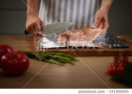 Person seasoning raw chicken breast with salt and spices on a cutting board, preparing a healthy meal with fresh ingredients Person seasoning raw chicken breast with salt and spices on a cutting board, preparing a healthy meal with fresh ingredients 141998005
