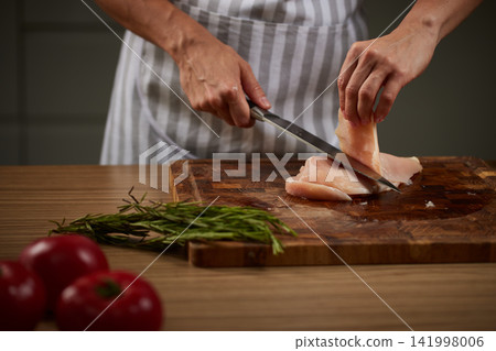 Person in an apron cutting fresh raw chicken breast with a knife on a wooden board, preparing healthy food Person in an apron cutting fresh raw chicken breast with a knife on a wooden board, preparing healthy food 141998006