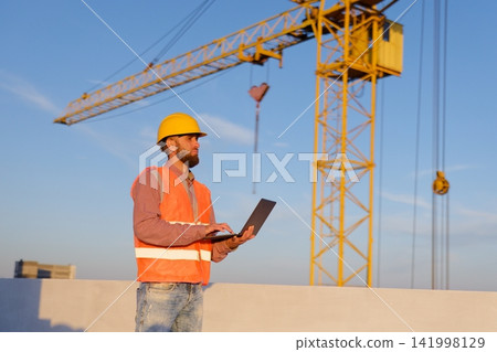 Construction engineer using laptop at building site with crane in background Construction engineer using laptop at building site with crane in background 141998129