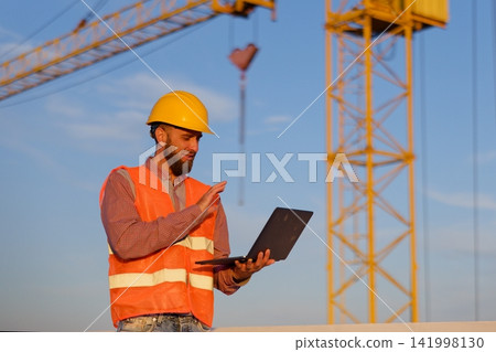 Construction engineer using laptop at building site with crane in background Construction engineer using laptop at building site with crane in background 141998130