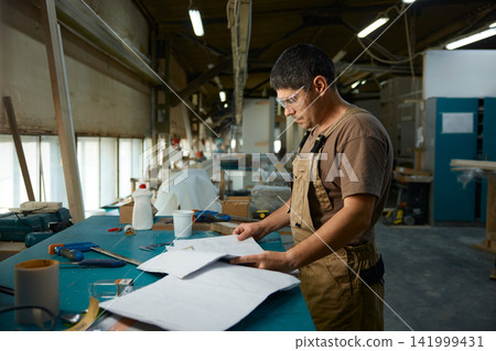 Experienced craftsperson analyzes complex design plans with surrounding tools and materials Experienced craftsperson analyzes complex design plans with surrounding tools and materials 141999431
