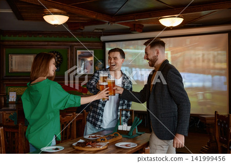 Friends cheer with beer glasses in cozy pub interior Friends cheer with beer glasses in cozy pub interior 141999456