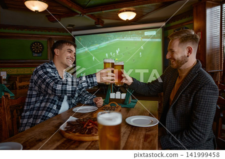Two white men clinking pints pub, colleagues celebrating goal while watching soccer on large screen Two white men clinking pints pub, colleagues celebrating goal while watching soccer on large screen 141999458