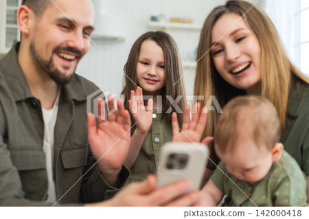 Happy family talk on video call with friends family. Mother father two children holding smartphone talking speaking online. Family mom dad parents baby girls kids smiling laughing enjoying moment Happy family talk on video call with friends family. Mother father two children holding smartphone talking speaking online. Family mom dad parents baby girls kids smiling laughing enjoying moment 142000418