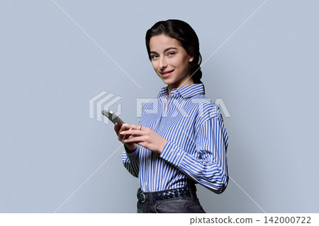 Young woman holding smartphone in half turn and looking at camera over grey background Young woman holding smartphone in half turn and looking at camera over grey background 142000722