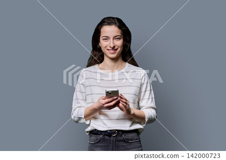 Smiling young woman holding smartphone and looking at camera over grey background Smiling young woman holding smartphone and looking at camera over grey background 142000723