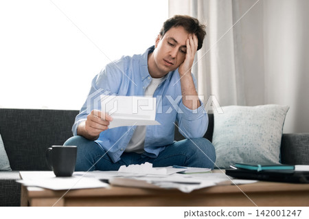 Stressed man reads overdue utility bills at home. He faces financial problems, rising energy prices, and high household expenses due to inflation and the economic crisis. Stressed man reads overdue utility bills at home. He faces financial problems, rising energy prices, and high household expenses due to inflation and the economic crisis. 142001247