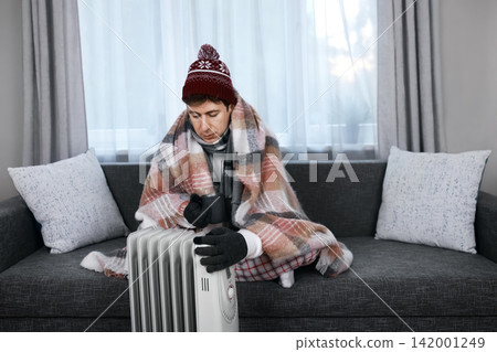 Freezing man wrapped in a blanket near a heater during a cold snap. He faces a home heating problem, weak heating, and rising energy costs due to the winter energy crisis. Freezing man wrapped in a blanket near a heater during a cold snap. He faces a home heating problem, weak heating, and rising energy costs due to the winter energy crisis. 142001249