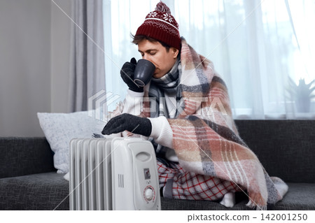 Man warms hands by a heater in cold weather. He drinks hot tea, facing a home heating problem, cold snap, weak heating, and rising energy costs amid an energy crisis. Man warms hands by a heater in cold weather. He drinks hot tea, facing a home heating problem, cold snap, weak heating, and rising energy costs amid an energy crisis. 142001250