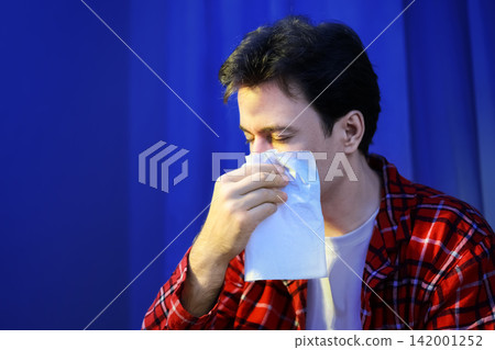 Sick man with flu symptoms uses a tissue for sneezing. Feeling unwell with a respiratory illness and cold infection, he needs home treatment and recovery. Sick man with flu symptoms uses a tissue for sneezing. Feeling unwell with a respiratory illness and cold infection, he needs home treatment and recovery. 142001252