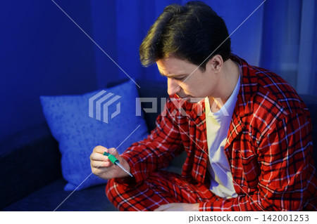 Sick man checks his fever with a thermometer. Feeling unwell with a seasonal cold and flu symptoms, he stays home for illness treatment, recovery, and fighting viral infection. Sick man checks his fever with a thermometer. Feeling unwell with a seasonal cold and flu symptoms, he stays home for illness treatment, recovery, and fighting viral infection. 142001253