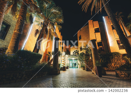 A courtyard at night, lined with illuminated stone buildings and palm trees. A courtyard at night, lined with illuminated stone buildings and palm trees. 142002236