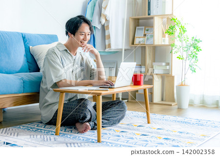 一位年輕男子身著便服,正在自家客廳工作。 一位年輕男子身著便服,正在自家客廳工作。 142002358
