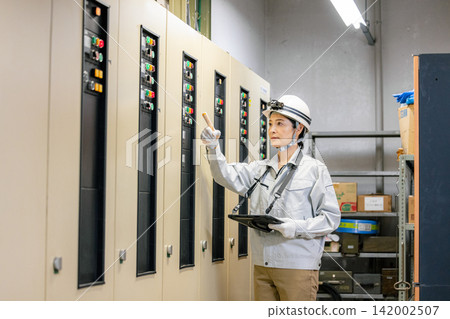 一名身穿工作服的女子手持平板電腦，站在電子控制面板前。 142002507