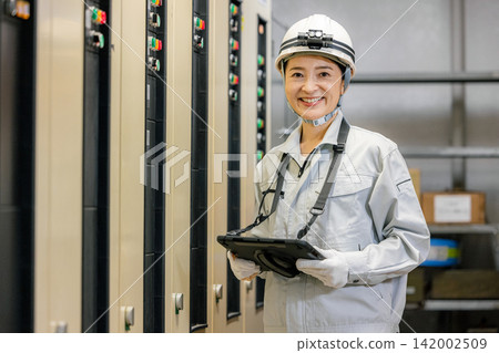 A woman in work clothes holding a tablet in front of an electronic control panel. A woman in work clothes holding a tablet in front of an electronic control panel. 142002509