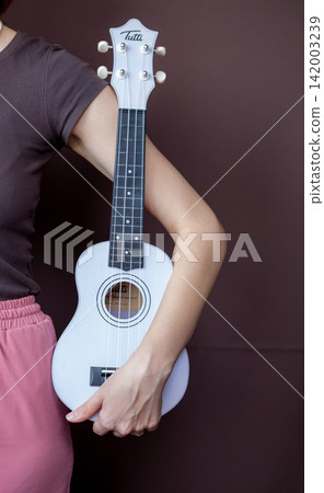 Woman's hand grips a white ukulele against a brown backdrop, highlighting the instrument's features and the person's casual attire Woman's hand grips a white ukulele against a brown backdrop, highlighting the instrument's features and the person's casual attire 142003239