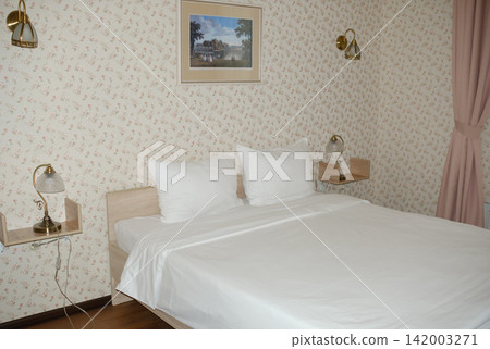 Modern bedroom features a double bed with white linens, decorative wall art above, and stylish bedside lamps against floral wallpaper 142003271