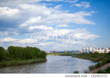 Scenic view of a river bordered by greenery, with a modern city skyline visible in the background under a partly cloudy sky Scenic view of a river bordered by greenery, with a modern city skyline visible in the background under a partly cloudy sky 142003272