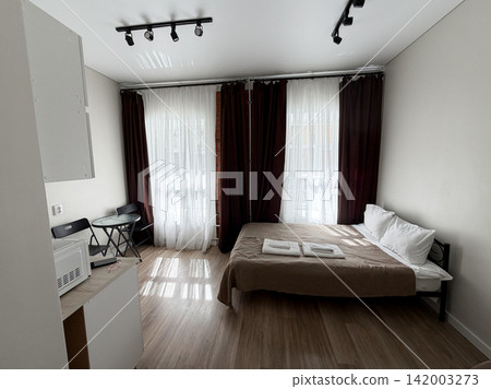 Modern bedroom interior showcases a bed with linens, curtains, and a small dining table with chairs, illuminated by natural light from windows 142003273