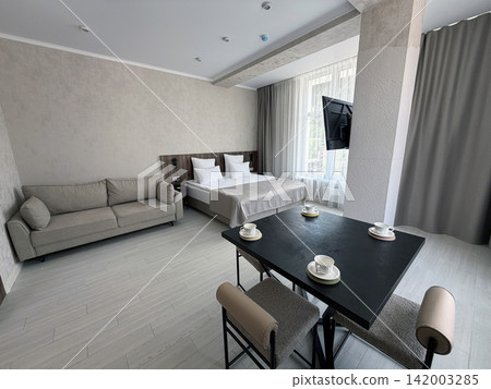 Modern hotel room showcases a double bed, sofa, dining table with chairs, and large windows allowing natural light to fill the area 142003285