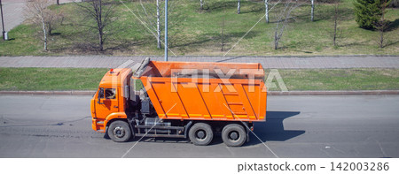 Orange dump truck carries soil along a city street, with green grass and trees lining the roadside in the background 142003286