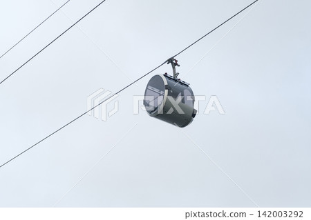 Aerial gondola cabin hangs from cables in the sky, highlighting contemporary transport design against a backdrop of gray clouds Aerial gondola cabin hangs from cables in the sky, highlighting contemporary transport design against a backdrop of gray clouds 142003292
