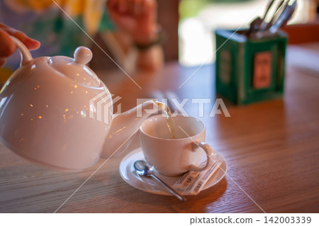 White teapot is pouring tea into a white cup placed on a wooden table, with a green container and spoon visible in the background White teapot is pouring tea into a white cup placed on a wooden table, with a green container and spoon visible in the background 142003339