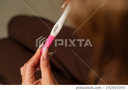 Woman examines a pregnancy test with a pink and white design while seated on a brown couch in a softly lit indoor environment Woman examines a pregnancy test with a pink and white design while seated on a brown couch in a softly lit indoor environment 142003343