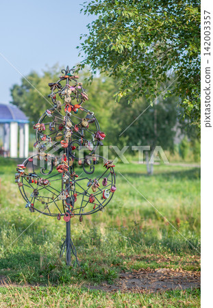 Metal tree sculpture decorated with various colorful locks stands in a park, surrounded by grass and trees under a bright blue sky Metal tree sculpture decorated with various colorful locks stands in a park, surrounded by grass and trees under a bright blue sky 142003357