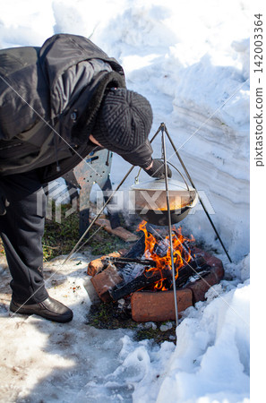Individual prepares food over an open fire in a snowy environment, utilizing a tripod to suspend a pot above the flames Individual prepares food over an open fire in a snowy environment, utilizing a tripod to suspend a pot above the flames 142003364