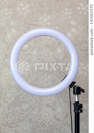 Circular LED ring light mounted on a black stand, positioned in front of a textured floral background, suitable for enhancing photography and video lighting Circular LED ring light mounted on a black stand, positioned in front of a textured floral background, suitable for enhancing photography and video lighting 142003375