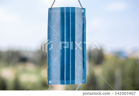 Blue disposable face mask suspended against a soft-focus outdoor backdrop featuring greenery and a bright sky, highlighting its design and texture Blue disposable face mask suspended against a soft-focus outdoor backdrop featuring greenery and a bright sky, highlighting its design and texture 142003407