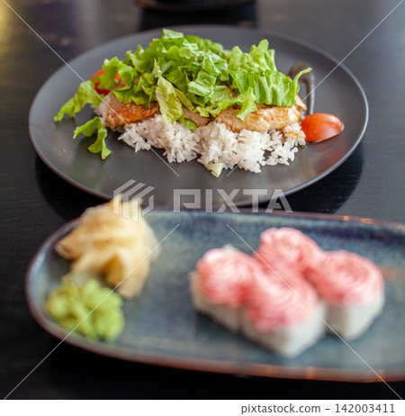 Sushi rolls with salmon and rice topped with fresh lettuce, accompanied by pickled ginger and wasabi on a dark wooden table setting Sushi rolls with salmon and rice topped with fresh lettuce, accompanied by pickled ginger and wasabi on a dark wooden table setting 142003411
