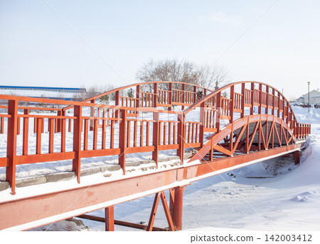 Red wooden bridge spans a snow-covered area, surrounded by winter scenery and distant structures under a clear blue sky Red wooden bridge spans a snow-covered area, surrounded by winter scenery and distant structures under a clear blue sky 142003412