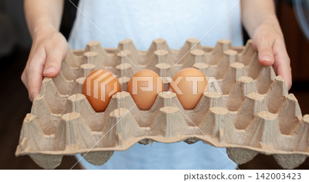 Individual presents a carton containing three brown eggs, showcasing the texture of the eggs and the egg carton in a kitchen setting Individual presents a carton containing three brown eggs, showcasing the texture of the eggs and the egg carton in a kitchen setting 142003423