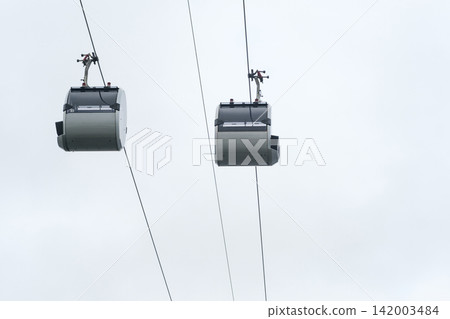 Two cable car gondolas are seen hanging from cables in a cloudy sky, representing a modern transportation system in an outdoor environment Two cable car gondolas are seen hanging from cables in a cloudy sky, representing a modern transportation system in an outdoor environment 142003484