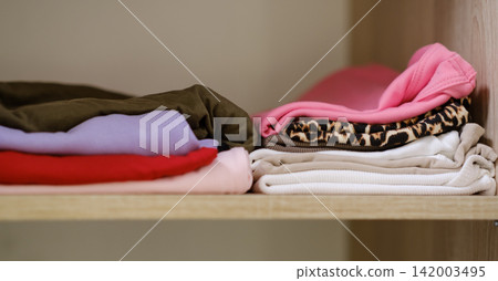 Neatly arranged clothing items in different colors and patterns stacked on a wooden shelf, highlighting organization and fabric variety Neatly arranged clothing items in different colors and patterns stacked on a wooden shelf, highlighting organization and fabric variety 142003495