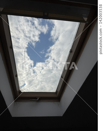 View of blue sky and white clouds through a skylight from inside a building, featuring a wooden frame and interior wall around the opening View of blue sky and white clouds through a skylight from inside a building, featuring a wooden frame and interior wall around the opening 142003538