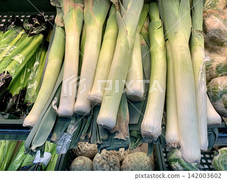 Fresh leeks are displayed prominently in a grocery store, with various vegetables and produce visible in the surrounding market environment 142003602