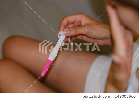 Woman sits on a bathroom floor holding a pregnancy test showing a positive result, dressed in a white dress, with a soft-focus background 142003666