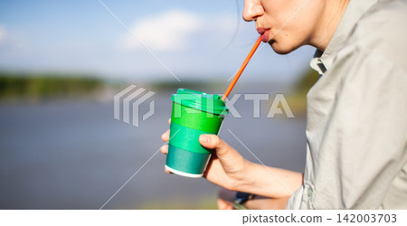 Woman sips from a green and blue disposable cup with a straw, standing by a river under a clear blue sky Woman sips from a green and blue disposable cup with a straw, standing by a river under a clear blue sky 142003703