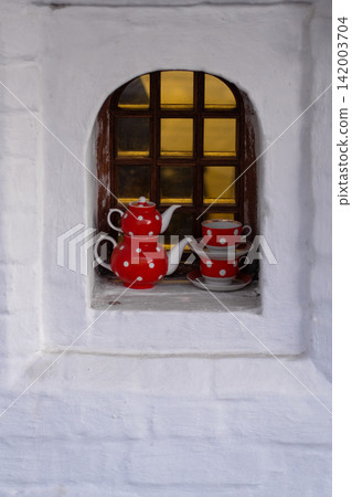 Red polka dot teapot and matching cups are arranged in a window with a wooden grid, set against a white wall background Red polka dot teapot and matching cups are arranged in a window with a wooden grid, set against a white wall background 142003704