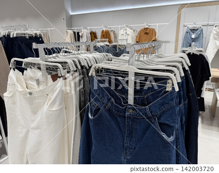 Assorted women's clothing items such as denim jeans and white pants are displayed on hangers in a contemporary retail store environment 142003720