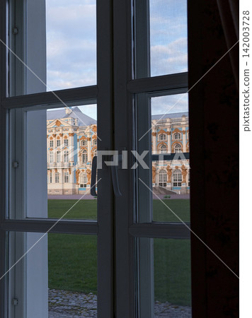 View of the Catherine Palace framed by an open window, highlighting its intricate architecture and green lawn in the foreground with clouds above View of the Catherine Palace framed by an open window, highlighting its intricate architecture and green lawn in the foreground with clouds above 142003728